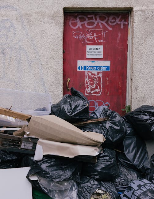 A large pile of black rubbish bags, some torn open, stacked against a beige building wall near a red metal door covered with graffiti. The bags vary in size and are placed on a pavement area, with additional debris including flattened cardboard boxes and a hospital-style rail. The door has several signs, one indicating 'No Money' and another with a blue 'Keep clear' notice, all partially obscured by the rubbish. The scene suggests an instance of waste accumulation awaiting collection, possibly related to an independent or private rubbish removal service such as Waste Removal Kentish Town, illustrating typical on-site clearance for domestic or commercial waste disposal outside standard council collection areas.