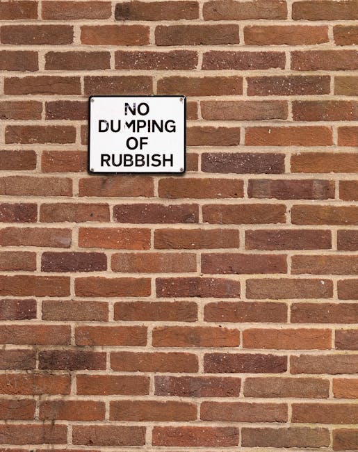 A rectangular metal sign mounted on a red brick wall, with the text 'NO DUMPING OF RUBBISH' printed in black capital letters on a white background. The sign's surface shows slight weathering, with some minor scratches and fading around the edges. The brick wall behind the sign features a traditional pattern with reddish-brown bricks and light-colored mortar joints, extending across the full visible background. The scene is evenly lit with natural light, casting minimal shadows, and the overall setting suggests an outdoor area where private rubbish removal or waste management services may be provided, aligned with objectives for independent waste handling and rubbish clearance.