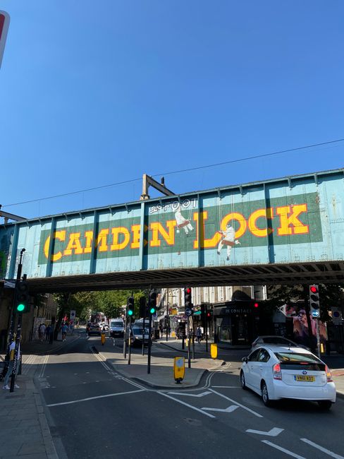 A view of a railway bridge overhead with a large, weathered sign displaying the words 'Camden Lock' in bold yellow letters against a teal background. The bridge is situated above a busy street intersection, where several vehicles, including a white car in the foreground, are navigating the roadway. Traffic lights are visible, with some showing green signals. Pedestrians can be seen on the sidewalks and crossing the street, some standing near the bus stop and others walking underneath the bridge. The sky above is clear and blue, providing natural daylight that highlights the industrial texture of the bridge's metal surface and the urban surroundings. This scene reflects an active part of Camden Town known for its markets and vibrant atmosphere, often associated with unique local businesses and independent collection of waste or rubbish, such as waste removal services provided by Waste Removal Kentish Town, in an area with a mix of commercial and pedestrian activity.
