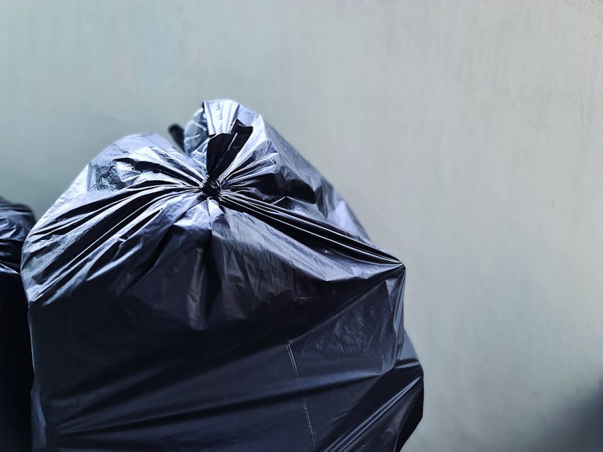 Close-up view of a tightly knotted black plastic rubbish bag made of shiny, smooth material, resting on a surface against a plain, light-colored wall. The bag's surface reflects ambient light, showing creases and folded areas, with some parts appearing more opaque and others slightly translucent. The bag is situated towards the lower left side of the frame, with other similar bags partially visible behind it. The environment suggests an indoor setting, possibly a storage or utility area used for waste collection or preparation for rubbish removal. The overall scene emphasizes the typical appearance of household rubbish ready for disposal, as managed by waste removal services like Waste Removal Kentish Town, supporting their focus on local rubbish clearance and alternative waste handling methods.