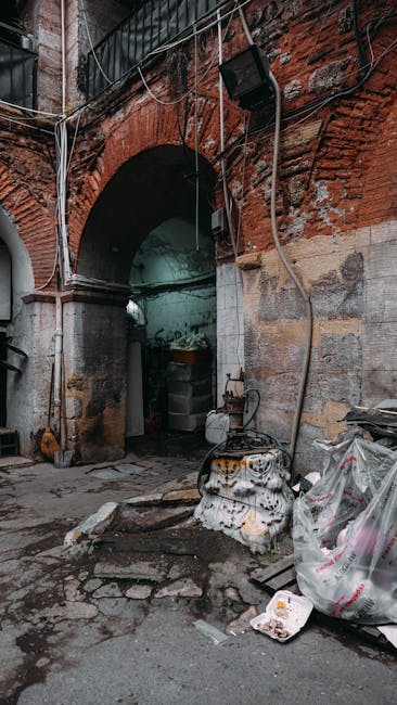 The image depicts a narrow alleyway or courtyard with an arched brick passageway leading into a shaded area. The surrounding brickwork appears aged, with some exposed and weathered bricks, and a section of the wall showing reddish-brown hues. Electrical wires run across the visible section of the wall and ceiling, connected to a black outdoor light fixture mounted on the brick arch. On the ground outside the arch, there are scattered pieces of rubbish, including a large, partially crumpled plastic bag filled with waste, some loose debris, and a metal container or utility box that appears worn and slightly stained. Nearby, an old, rusted, and possibly discarded appliance or part of a mechanical setup is visible, with signs of rust and dirt. The overall atmosphere suggests a neglected urban environment, where waste has accumulated near the entrance, exemplifying typical scenes where private waste disposal services, like those offered by Waste Removal Kentish Town, might be called upon for clearance or rubbish removal in such settings.