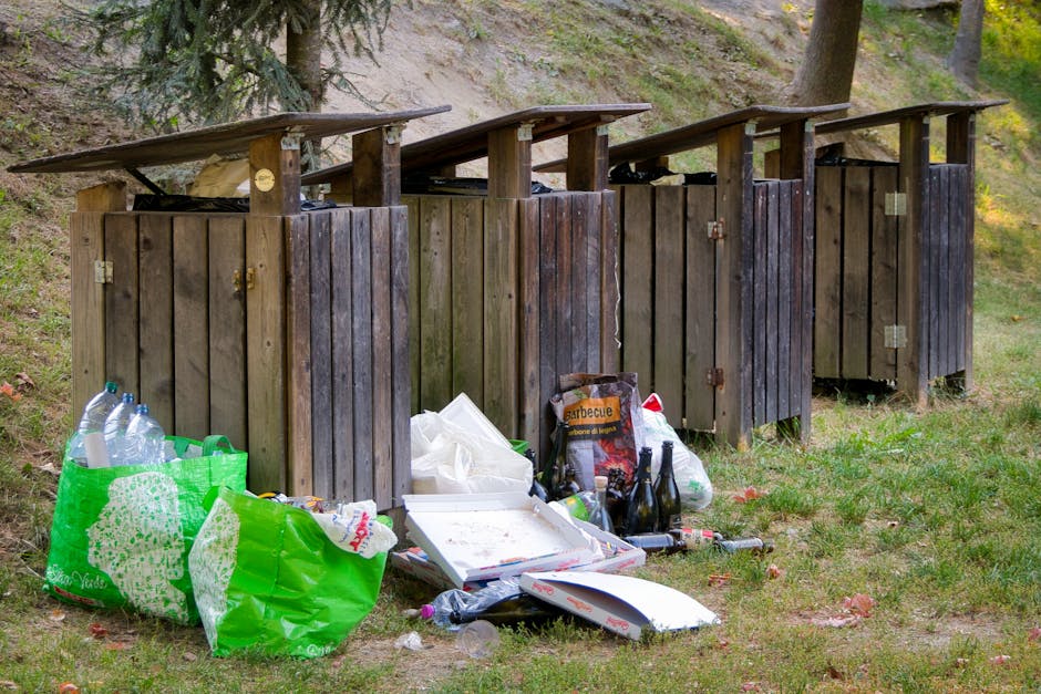 A row of four wooden outdoor toilets with sloped roofs, constructed from weathered vertical wooden planks in varying shades of brown, are situated on a grassy area with a slight incline, bordered by trees and a dirt embankment in the background. In front of the toilets, litter has accumulated, including a large green plastic bag filled with empty plastic bottles, a white pizza box with remnants of food, a partially crushed black cardboard box labeled 'barbecue', and several glass bottles, some of which are broken, along with a clear plastic bag containing more waste. Loose items, such as plastic bottles, a crumpled paper bag, and scattered trash, are strewn across the grass near the waste, indicating a collection of improperly disposed rubbish. The scene appears to be outdoors in a park or woodland area, with natural light illuminating the scene and casting slight shadows, suggesting daytime. The presence of the waste reinforces the importance of proper rubbish removal services to prevent environmental littering in scenic outdoor locations. Waste Removal Kentish Town handles such rubbish clearance to maintain cleanliness and natural beauty.