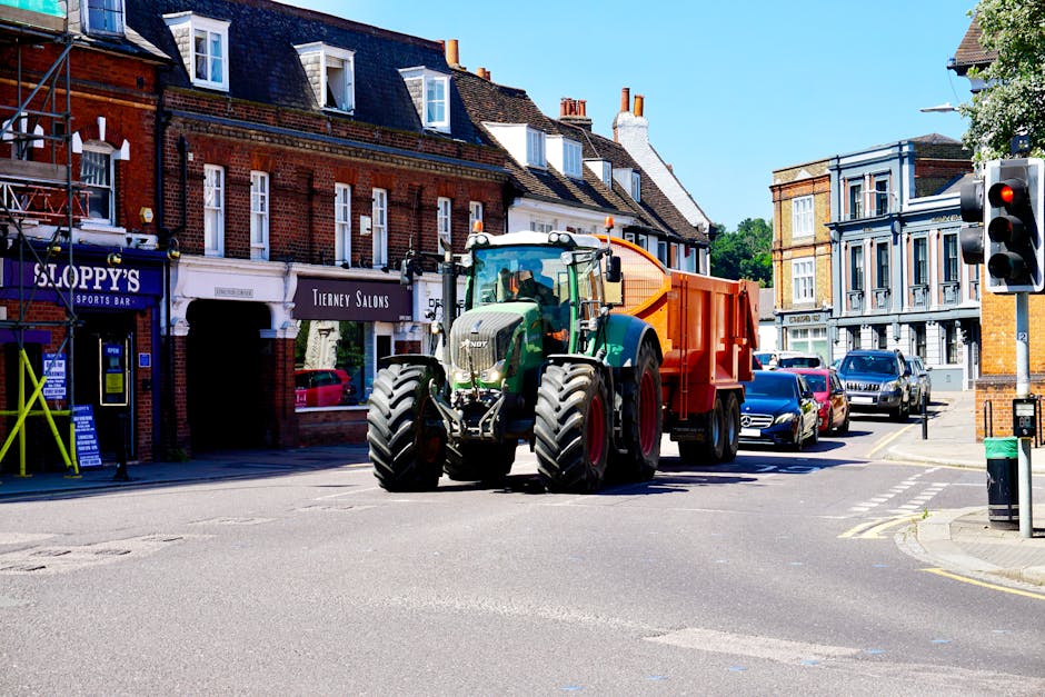 A large green tractor with black tires and a white cab is positioned at the intersection of a residential street, pulling an orange waste container on its back. The setting includes multi-storey brick and stucco buildings on either side of the street, with retail shops visible on the left, such as a sports bar and a salon. Several parked cars line the street in the background, and a traffic light is visible on the right side of the image, showing a red signal. The scene appears to be during daytime with clear blue skies, providing natural lighting that highlights the textures of the brick facades, the smooth surfaces of the vehicles, and the reflective finish of the waste container. The image illustrates an example of private waste management or rubbish removal service, with the tractor likely involved in on-site waste collection or clearance, supported by Waste Removal Kentish Town.