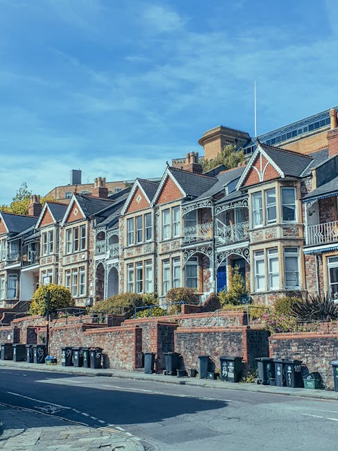 A row of Victorian-style terraced houses with ornate white ironwork balcony railings and bay windows, constructed from red brick with decorative gable features and slate roofs. The houses are set back from the street, with small front gardens enclosed by low brick walls and paved steps leading up to the front doors. The street in the foreground features a paved pavement and a line of black wheelie bins positioned along the curb, indicating waste collection activities. The sky above is clear and blue, suggesting a bright day. The scene is situated in an urban residential area, with the houses showing signs of well-maintained facades. The presence of waste bins implies that the area may utilize private rubbish collection services, as offered by Waste Removal Kentish Town. The environment appears quiet and orderly, with natural sunlight illuminating the buildings and street scene.