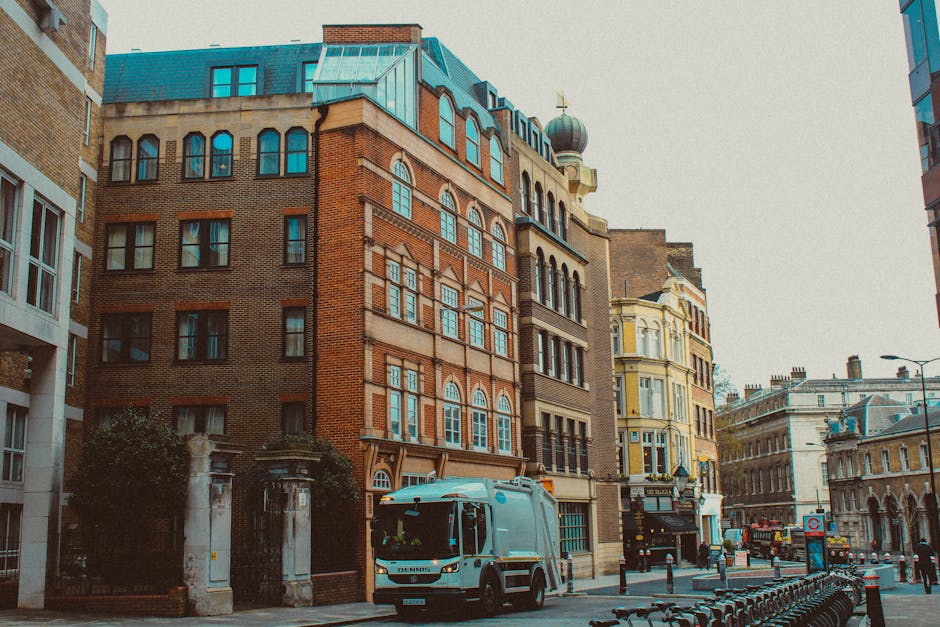 A street scene featuring a row of multi-storey buildings with varied architectural styles, including brick facades and ornate window detailing, situated in an urban environment. In the foreground, a waste collection vehicle with a silver and blue finish is parked along the curb, adjacent to a series of metal bike racks. The street is lined with bollards and is illuminated by natural daylight, with no visible pedestrians or other vehicles. To the left, part of a white modern building can be seen, while on the right, a combination of historic and contemporary structures extends into the background. The scene suggests an area where private waste removal services, such as those provided by Waste Removal Kentish Town, are utilized for managing rubbish in a densely built district, supporting the context of alternative waste handling methods outside of municipal collection on busy city streets.
