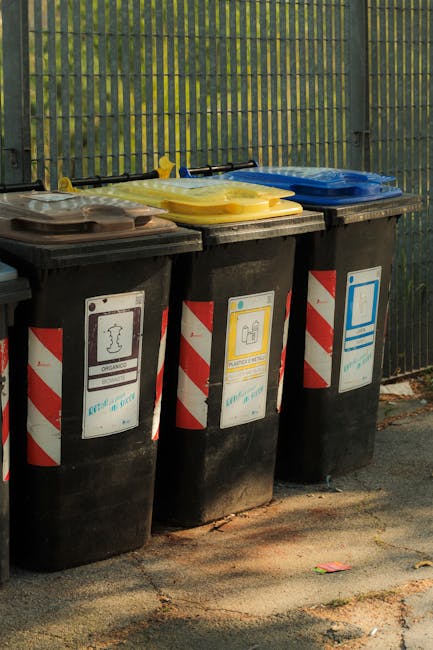 Three large black wheelie bins are positioned side by side on a paved outdoor surface, each fitted with a differently coloured lid: the left bin has a dark grey lid, the middle bin has a yellow lid, and the right bin has a blue lid. The bins are made of sturdy plastic with a textured surface and are marked with labels featuring icons and striped red and white hazard patterns, indicating their use for recycling or waste disposal. The lids are flat with slightly raised edges, and appear to be made of smooth, durable plastic. Behind the bins, there is a tall, green metal fence with vertical slats secured by horizontal supports, providing boundary fencing for the waste collection area. The background shows a blurred, leafy environment suggesting an outdoor setting. The scene is illuminated by natural daylight, casting subtle shadows on the asphalt pavement, which displays small cracks and scattered debris, characteristic of an urban or suburban waste management zone. Waste Removal Kentish Town's service for rubbish disposal is demonstrated in the context of organized waste separation for private disposal outside regulated council schemes.