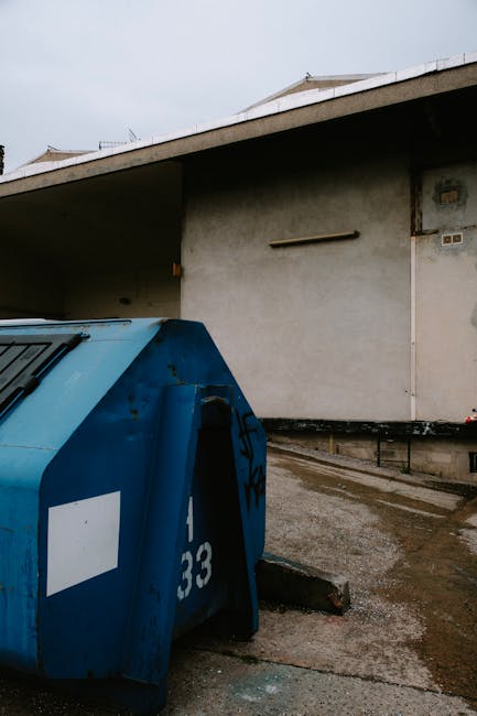 A large, blue, wheeled rubbish bin with a curved lid, showing visible signs of dirt and scratches on its surface, is positioned on a concrete pavement in front of a plain, light gray building wall with minimal detailing. The bin has a white rectangular label on the side and the number '33' painted in white near the bottom. The surrounding environment appears to be an urban or commercial area, with wet pavement indicating recent rain. In the background, part of the building's roof and a small dark metal railing are visible along the edge of the pavement. The scene is illuminated by natural daylight, with overcast skies giving a diffuse light, underscoring the functional, no-frills nature of the waste collection area. This setting exemplifies a typical private waste handling space, often managed by waste removal services like Waste Removal Kentish Town, emphasizing on-site rubbish storage designated for independent collection purposes in line with non-local authority disposal practices.