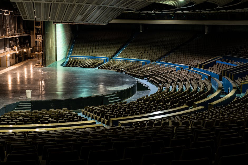 An empty indoor theatre or concert hall featuring a large, curved stage with a smooth, dark surface and steps leading up to it, surrounded by tiered seating arranged in a semi-circular layout. The seats are upholstered in dark brown fabric and divided by light blue and yellow safety barriers. On the left side, there are industrial-style metal shelving units and stage lighting fixtures along the backstage area, with warm, focused lighting illuminating the space and creating a contrast between the dark seating and the brightly lit stage area. The overall environment appears clean and well-maintained, with no visible clutter or objects, indicating it is set up for a performance or event and possibly prepared for cleaning or maintenance using specialist waste removal services, as inferred from the context of debris or waste management related to event venues.