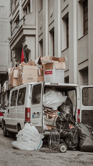 A white van with its rear doors open, parked along a city street adjacent to multi-storey buildings with beige facades and rectangular windows. The van's interior is filled with various waste materials, including large black and white rubbish bags, flattened cardboard boxes, and other miscellaneous refuse. Several cardboard boxes, some partially crushed and others intact, are stacked on top of the van, extending above the roofline. A small hand trolley loaded with additional black rubbish bags is positioned on the pavement near the rear of the van, suggesting the process of waste collection or loading. The surroundings include a narrow street with a pavement on the right side, and the area appears to be part of an urban setting, potentially representing an example of private waste handling or an independent rubbish clearance operation facilitated by Waste Removal Kentish Town. Natural daylight provides soft shadows, highlighting the textures of the cardboard, plastic bags, and the building surfaces. The scene visually captures a typical rubbish removal activity involving the sorting and loading of waste materials for disposal.