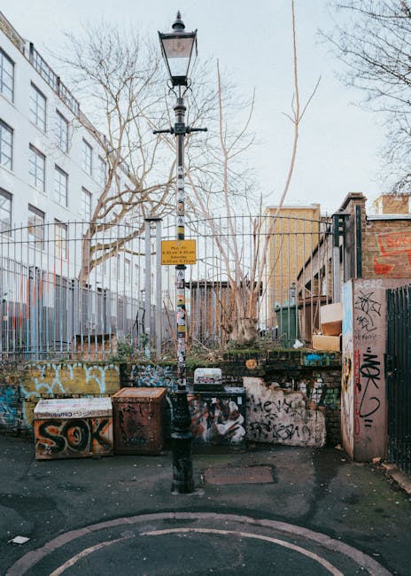 The image depicts a small urban outdoor area located on a paved surface, possibly a back alley or courtyard, with a tall, black, vintage-style lamppost positioned centrally. Attached to the lamppost is a yellow warning or informational sign. Surrounding the area are graffiti-covered concrete structures and low walls, with visible tags and spray paint in various colors, predominantly black and orange. In the background, there are metal fences, some with additional graffiti, enclosing what appears to be a small garden or storage area filled with leafless trees and additional urban debris. To the right, part of a brick wall and a green metal fence are visible, with some cardboard boxes and other discarded items stacked nearby. The overall scene appears to be in a neglected, public space affected by graffiti and litter, reflecting common urban waste collection and rubbish disposal challenges, possibly indicating a need for private rubbish removal or on-site clearance services provided by Waste Removal Kentish Town.