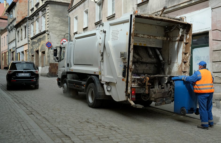 A white commercial rubbish collection truck is parked on a cobblestone street in an urban area, with its rear compartment open to reveal a rusted metal interior designed for waste loading. The truck's surface is slightly weathered, with visible signs of use and some rust on the edges of the open compartment. To the right, a worker dressed in a high-visibility orange vest, blue uniform, and protective gloves is positioned beside a blue wheeled bin, actively loading the bin into the truck's container. The scene is set against a backdrop of old, multi-storey buildings with a mix of stone and plaster facades, some showing signs of wear and peeling paint. There is minimal street traffic, with a black car parked further down the road, and the overall lighting suggests overcast or diffused natural daylight, creating a neutral atmosphere typical of a routine waste collection operation, indicative of private waste handling and alternative rubbish removal services in urban environments.