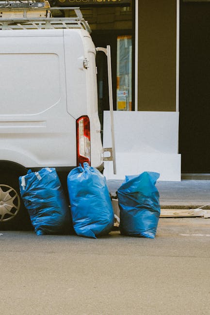 The image shows the rear side of a white commercial van parked on a pavement, with three large blue plastic rubbish bags placed directly on the ground next to it. The bags are filled with waste, appearing bulky and slightly crinkled, with varying shades of blue. The van has a partially visible rear door, with a vertical red and white indicator light on the left side. In the background, a building features a window and a white barrier or step, possibly part of a shopfront or commercial premises. The environment appears clean and well-maintained, with the bags positioned on a smooth asphalt surface. This scene illustrates an example of private waste collection or on-site rubbish removal, consistent with services offered by companies like Waste Removal Kentish Town, emphasizing efficient and discreet rubbish disposal options.