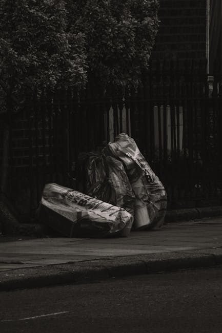 Three large black plastic garbage bags filled with rubbish are resting on the edge of a pavement in front of a black metal fence with vertical bars. Behind the fence, there is dense green foliage and a tall wooden structure, possibly a shed or fence panel. The scene appears to be outdoors during daylight, with some shadows cast by nearby objects. The rubbish bags are slightly crinkled and appear to be tightly packed, suggesting they contain household waste or garden debris. The environment indicates an area where private waste disposal occurs, with Waste Removal Kentish Town offering alternative rubbish collection services that might include on-site clearance or scheduled collections for bulky waste. The image emphasizes the importance of responsible disposal of rubbish, especially in urban settings, highlighting the need for reliable waste management solutions. The overall scene is professional and straightforward, showing typical rubbish accumulation in a residential or shared outdoor space, relevant to services specializing in rubbish removal and waste handling.